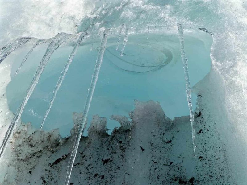 A close up of ice hanging in long stalactites from a glacier. To the bottom of the frame the ice has speckles of dark volcanic ash in it, contrasting with the blue reflection of the sky.