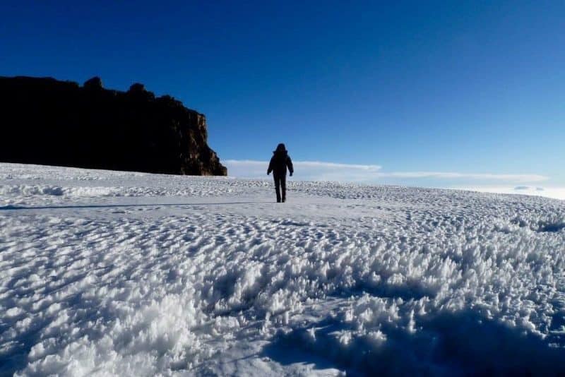 A person walking across a vast glacier at crater camp Kilimanjaro with a big rocky outcrop to the left of the frame and blue sky and bright sunshine in the background.