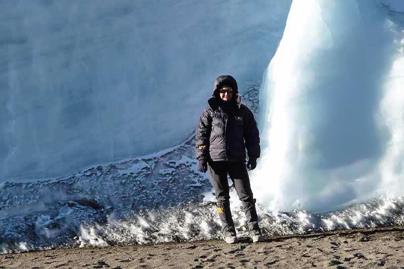 A person dressed in a black down jacket with a hood, black hiking trousers and gaiters standing in front of a large white glacier in bright sunshine. Volcanic soil at the base of the large ice wall and under the person's feet.