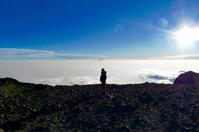 A person standing in silhouette on the edge of Kilimanjaro's Western Breach. Above the clouds, the landscape is rocky and barren. The background is bright sunshine with a blue sky.