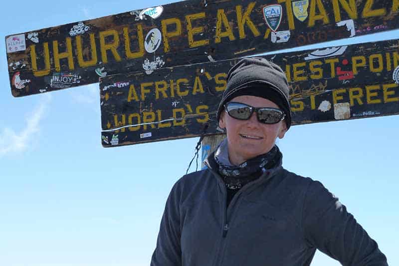 A person dressed in a gray fleece jacket and a beanie hat with sunglasses on stands in front of the summit sign at the top of Kilimanjaro. The background is blue sky in bright sunshine.