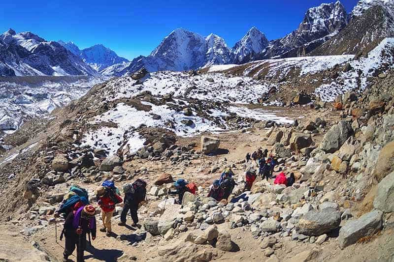 A horizontal image of a group of people backpacking in Nepal pictured on a backdrop of mountains and blue sky.