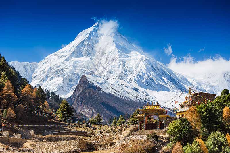 A horizontal image of monasteries in a Nepalese village surrounded by snow capped peaks and alpine vegetation.