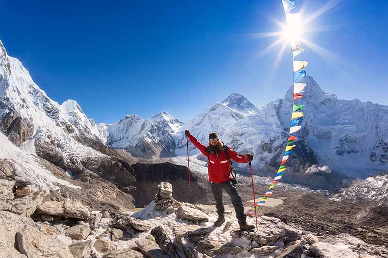 A horizontal image of a man carrying trekking poles pictured with prayer flags and mountains in the background.