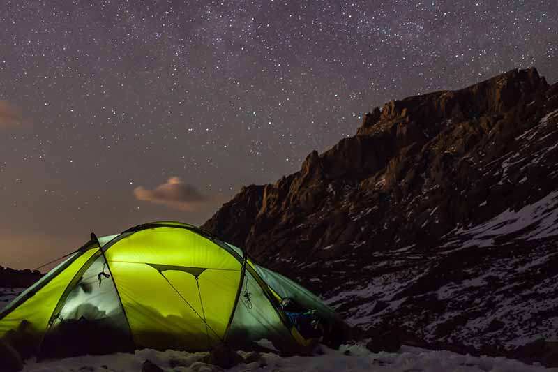 A green illuminated dome tent set up at the base of a mountain in a lightly snowy landscape at night. The background is dark ridges of the hill, and a starry night sky.